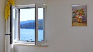 A room with a window overlooking a body of water and hills, a painting of flowers on the wall, and a curtain partially drawn. - Stunning home in Rabac (Rabac)
