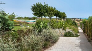 Ein Kiesweg führt zu einem Poolbereich, der von üppiger Vegetation und Kakteen umgeben ist, mit freiem Blick auf das Meer im Hintergrund.