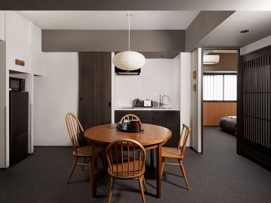 Uma sala de jantar moderna com uma mesa redonda de madeira, quatro cadeiras de madeira, um candeeiro pendente e uma cozinha ao fundo.