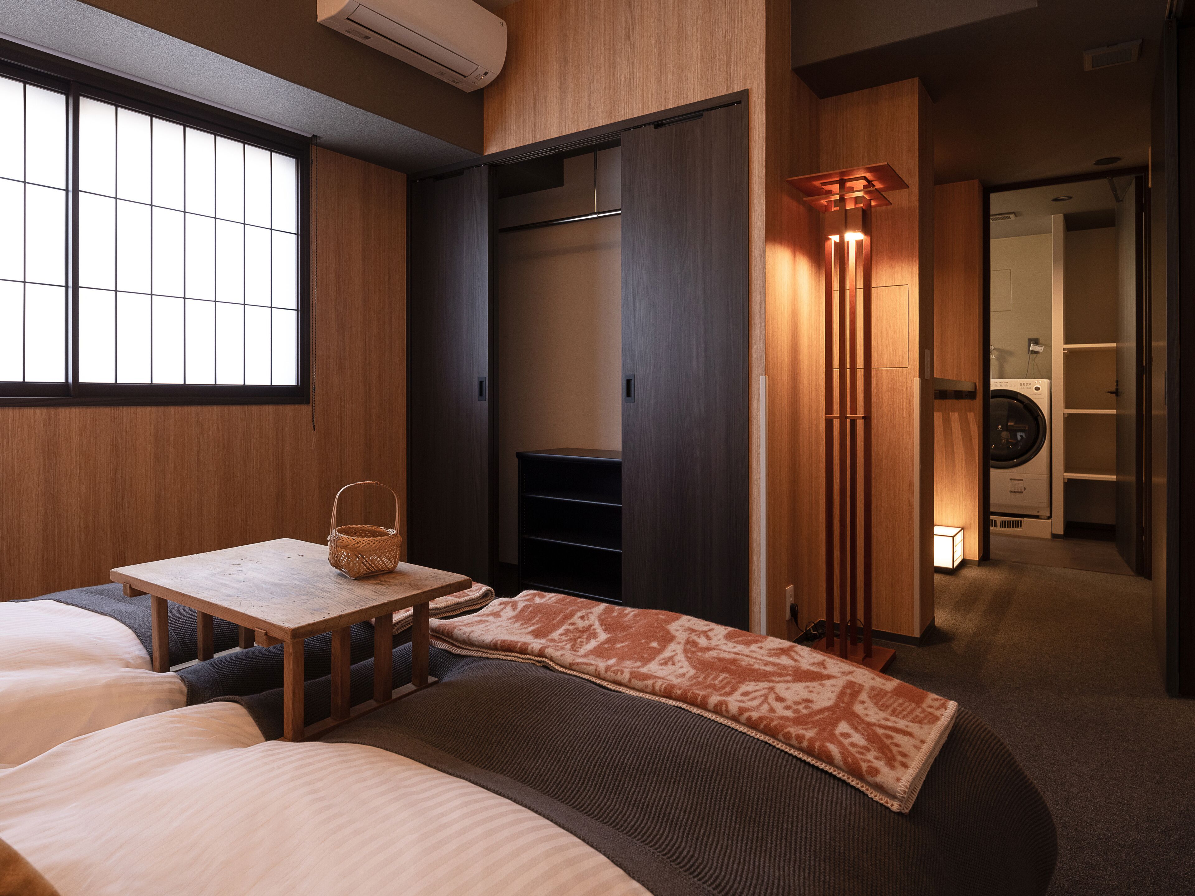 A hotel room with a bed, a small table with a basket, a wardrobe, and a washing machine in the background.