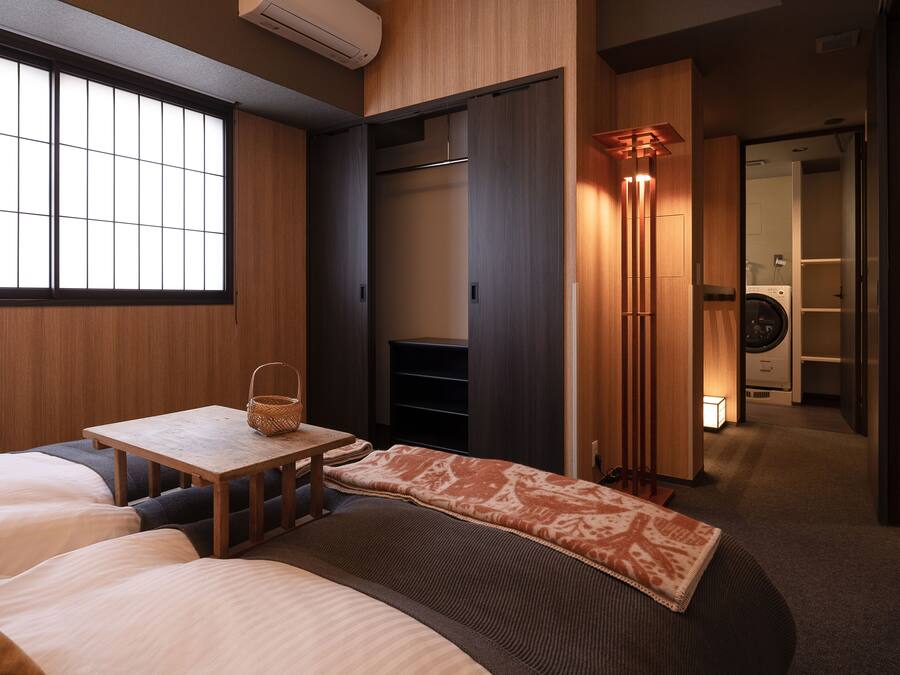A hotel room with a bed, a small table with a basket, a wardrobe, and a washing machine in the background.
