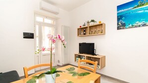 Ein Zimmer mit Esstisch aus Holz, Stühlen, einer Zimmerpflanze, einem Fernseher und einem Regal mit verschiedenen Gegenständen.