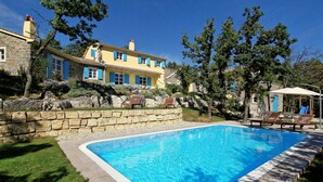 Une piscine bordée d’un mur en pierre et entourée de chaises longues, le tout adossé à une maison en pierre aux volets bleus.