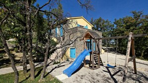 Un terrain de jeux avec un toboggan bleu, une balançoire en bois et un bâtiment en pierre en arrière-plan.