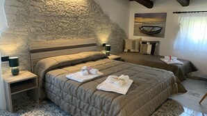 Une chambre avec un mur en pierre, un lit recouvert d’une courtepointe beige, deux tables de chevet avec des lampes et un tableau représentant un bateau accroché au mur.