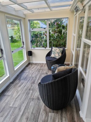 Ein sonnendurchflutetes Zimmer mit zwei Korbstühlen und einem kleinen Tisch, großen Fenstern mit Blick auf Grünflächen und einem Deckenventilator.