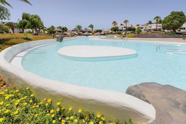 A swimming pool with a circular island, surrounded by a landscaped area with rocks and yellow flowers. - Lanzahost "Casa Christina" (Costa Teguise)