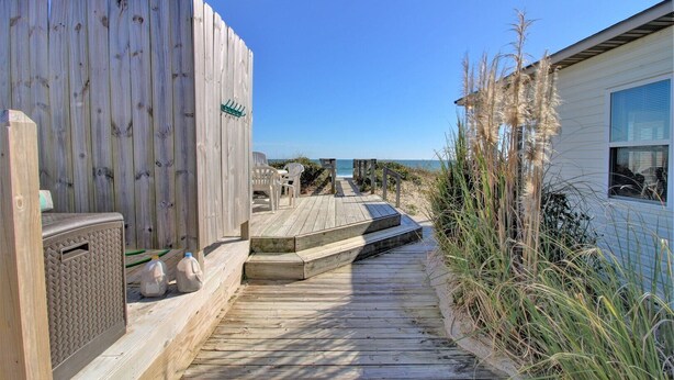 A wooden deck leading to a beach area with a white fence, a small outdoor seating area, and a house with a window. - Wave To Cool: 4-bedroom oceanfront cottage in Surf City, NC. (Surf City)