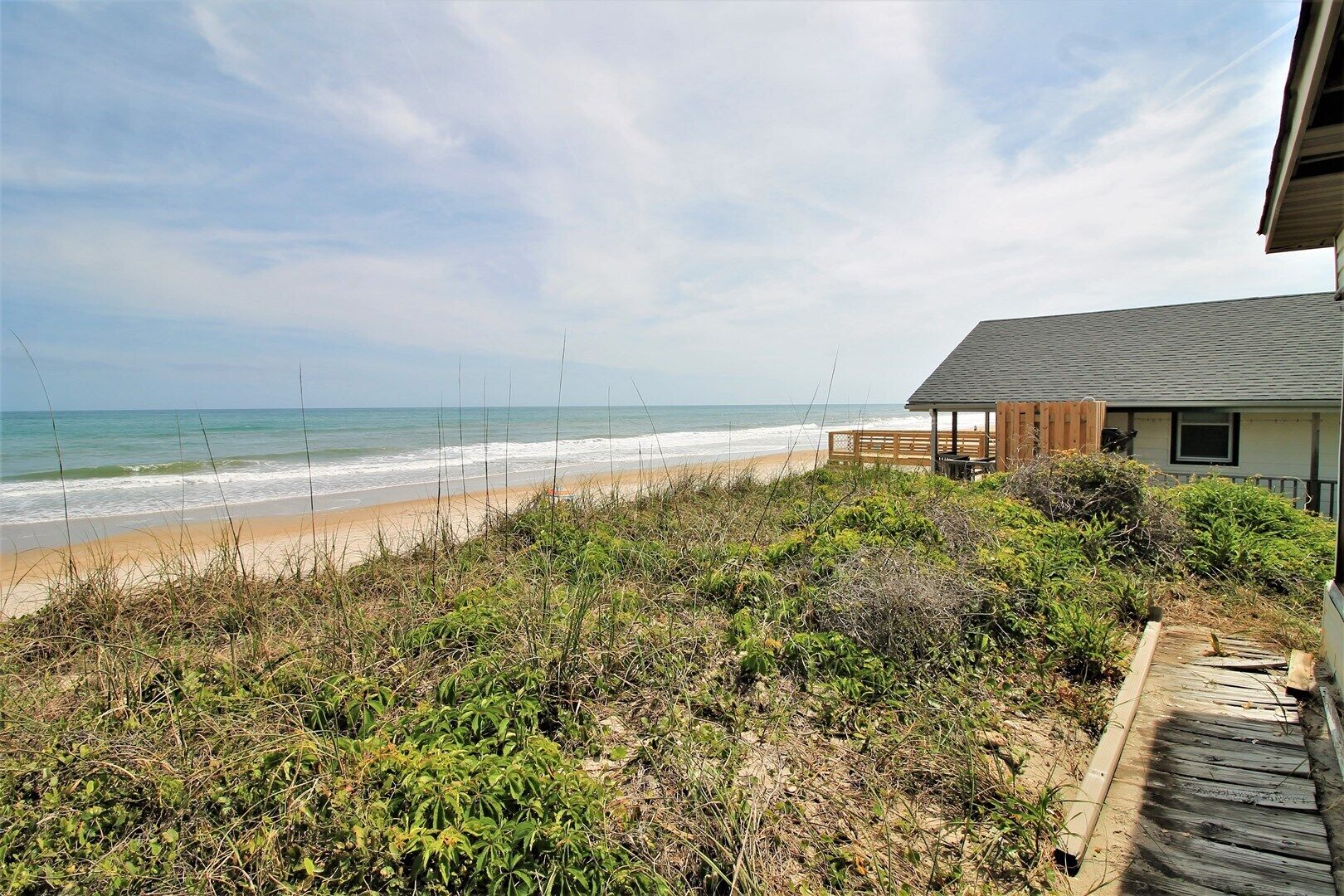 A beach view with a wooden deck leading to a house with a grey roof.