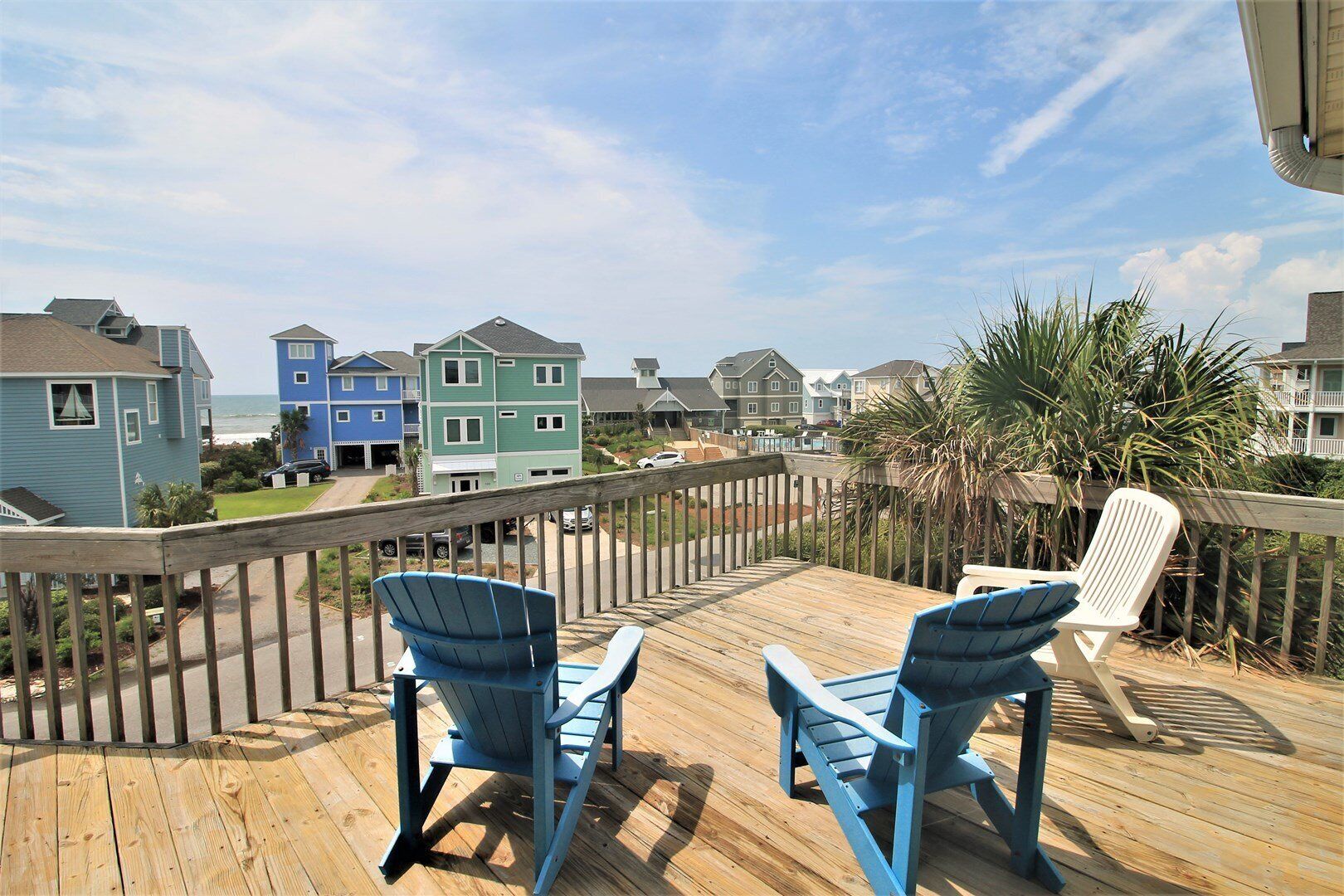 A wooden deck with blue Adirondack chairs and a white chair, overlooking a row of colorful houses by the sea.