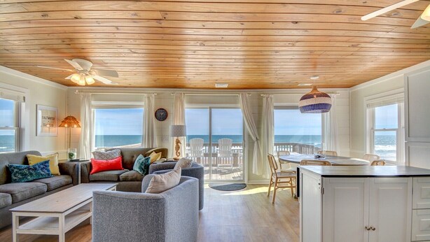 A spacious living room with a wooden ceiling, large windows overlooking the ocean, a dining area with wicker chairs, and a kitchen with white cabinets. - Oceanfront, 3-bedroom Whale Song in Surf City - perfect for 7 guests. (Surf City)