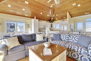 A spacious living area with a wooden ceiling, a large sofa set, a coffee table, and a kitchen in the background.