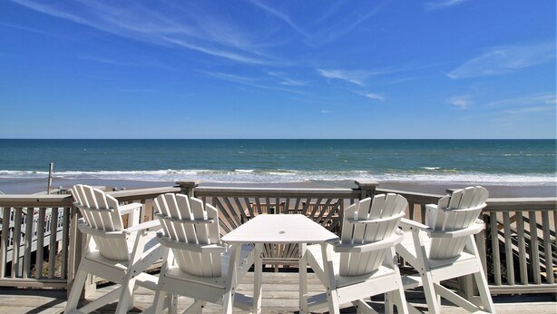 A wooden deck with white Adirondack chairs and a table overlooking the ocean. - C-Breeze: Oceanfront 4BR home in Surf City, NC, sleeps 13 comfortably. (Surf City)