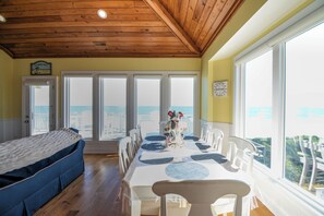 A room with a wooden ceiling, a dining table with chairs, a sofa, and large windows offering a view of the sea.