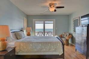 A bedroom with a bed, a dresser, a ceiling fan, a lamp, and a view of the ocean through a door.