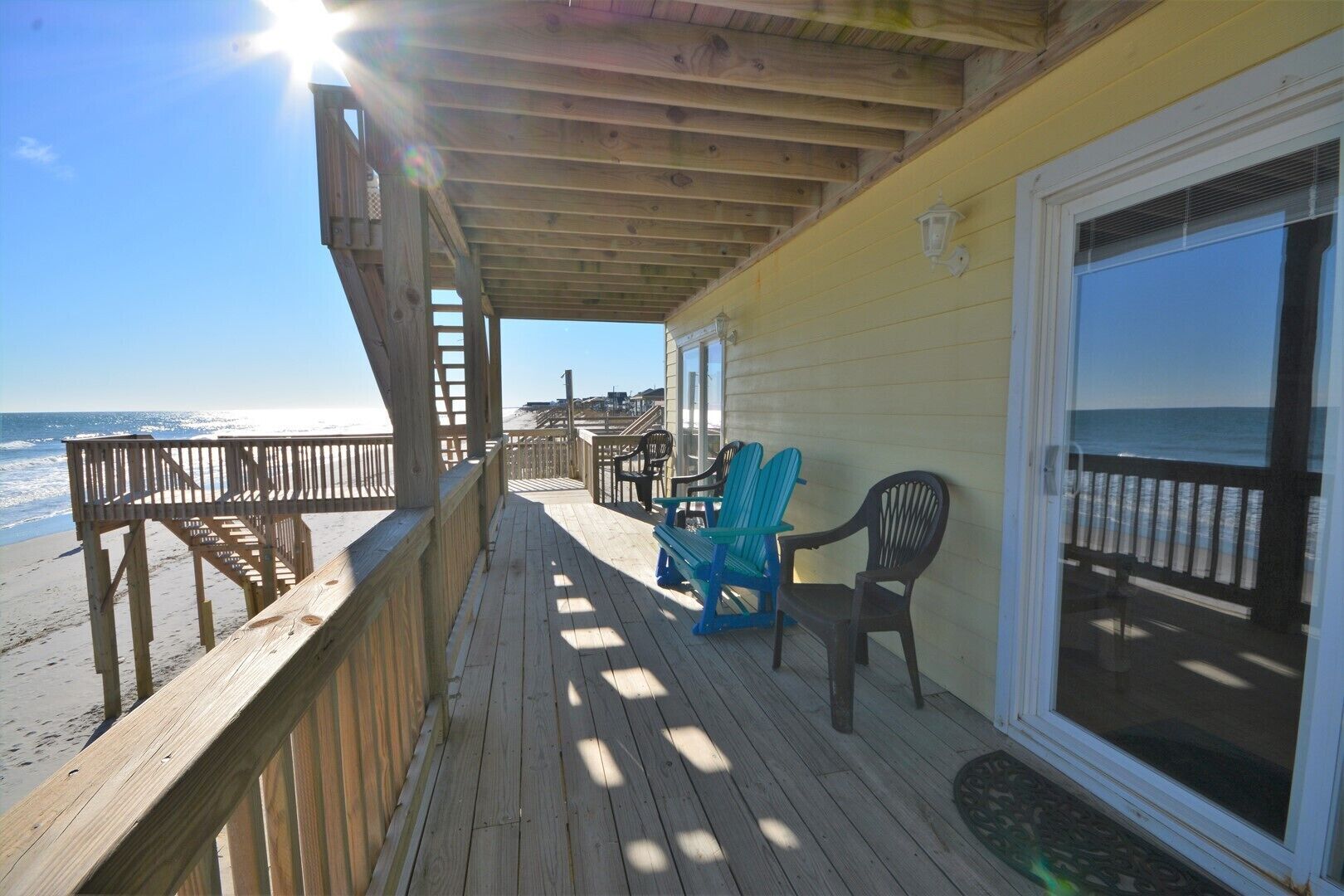 A wooden deck with a view of the beach, a glass door, and outdoor seating.