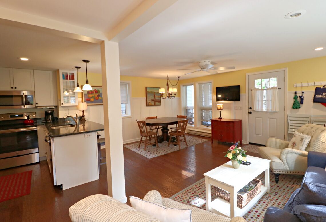 A modern kitchen with stainless steel appliances, a dining area with a round table and chairs, a living room with a sofa and a coffee table, and a TV mounted on the wall.