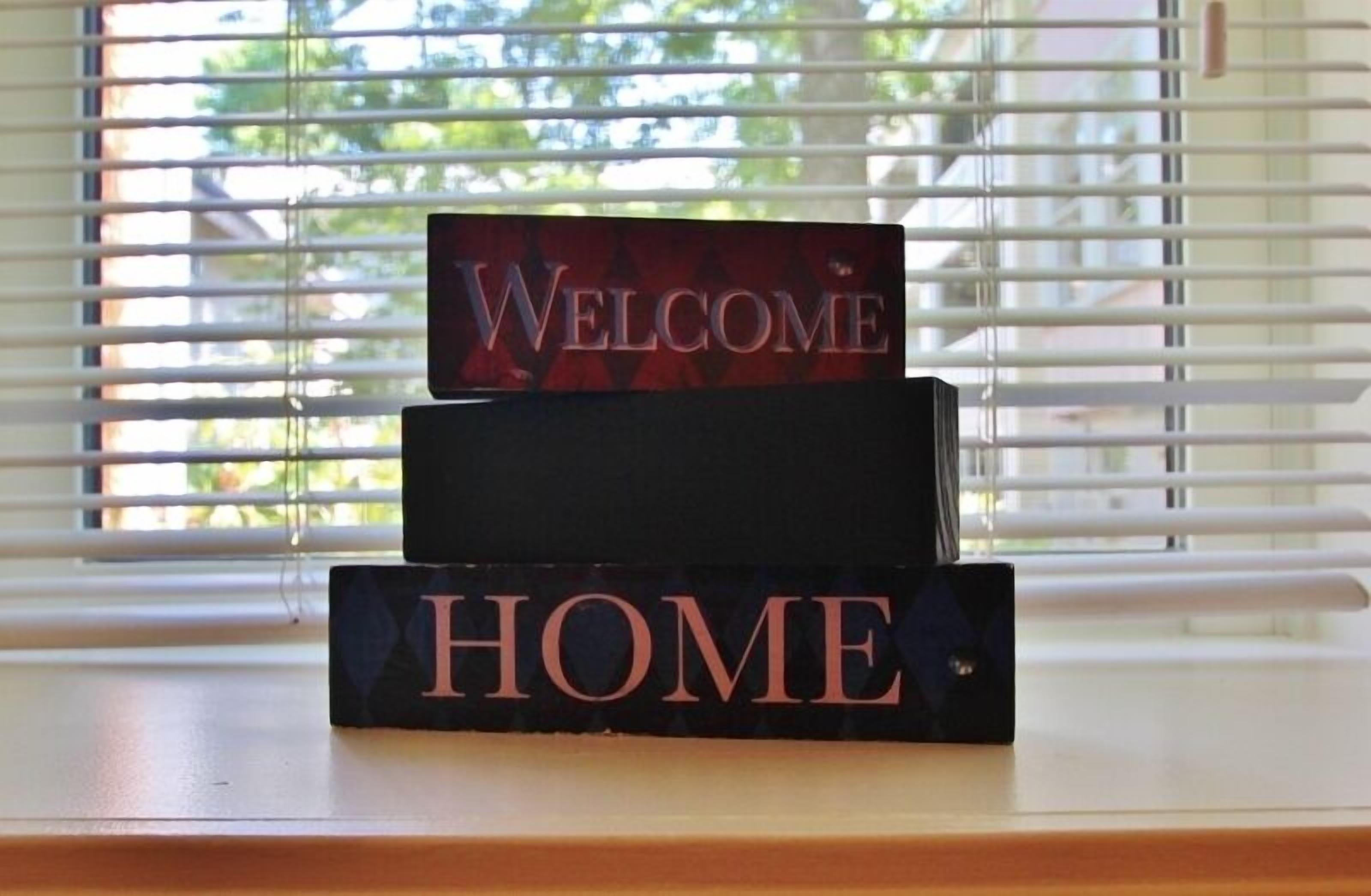 Een welkomstbord met het woord 'WELCOME' bovenaan en 'HOME' eronder, geplaatst op een vensterbank met zonwering op de achtergrond.