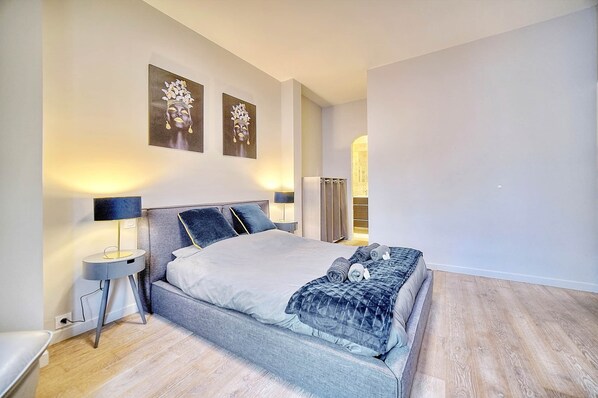 Une chambre moderne avec un grand lit, deux œuvres d’art accrochées au mur, une table de chevet avec une lampe, et une vue sur le paysage urbain par la fenêtre.