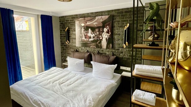 A hotel room with a large bed, a brick wall, a painting, a shelf with towels, and a window with blue curtains. - IQ Hotel (Hannover)