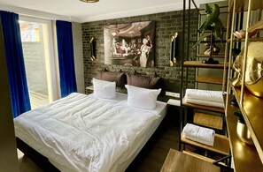 Habitación de hotel con una cama grande, pared de ladrillo, un cuadro, estante con toallas y ventana con cortinas azules.