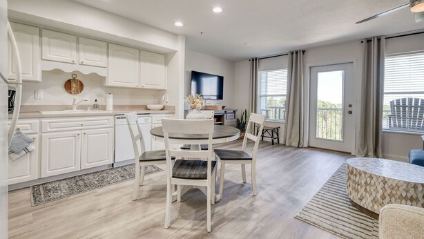 A modern kitchen with white cabinetry, a dining area with a round table and chairs, and a balcony with a view. - Nature's Landing B203 Seasalt in Historic CedarKey (Cedar Key)