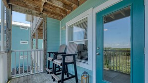 A covered porch with two Adirondack chairs, a wooden deck, and a teal door with a window. - Nature's Landing B203 Seasalt in Historic CedarKey (Cedar Key)