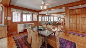 A spacious dining area with a glass table and wicker chairs, wooden walls, and large windows with blinds. - Spencer's Landing - Ocracoke Island (Ocracoke)