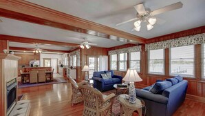A spacious living room with a fireplace, blue sofas, wicker chairs, and a dining area with a kitchen in the background. - Spencer's Landing - Ocracoke Island (Ocracoke)