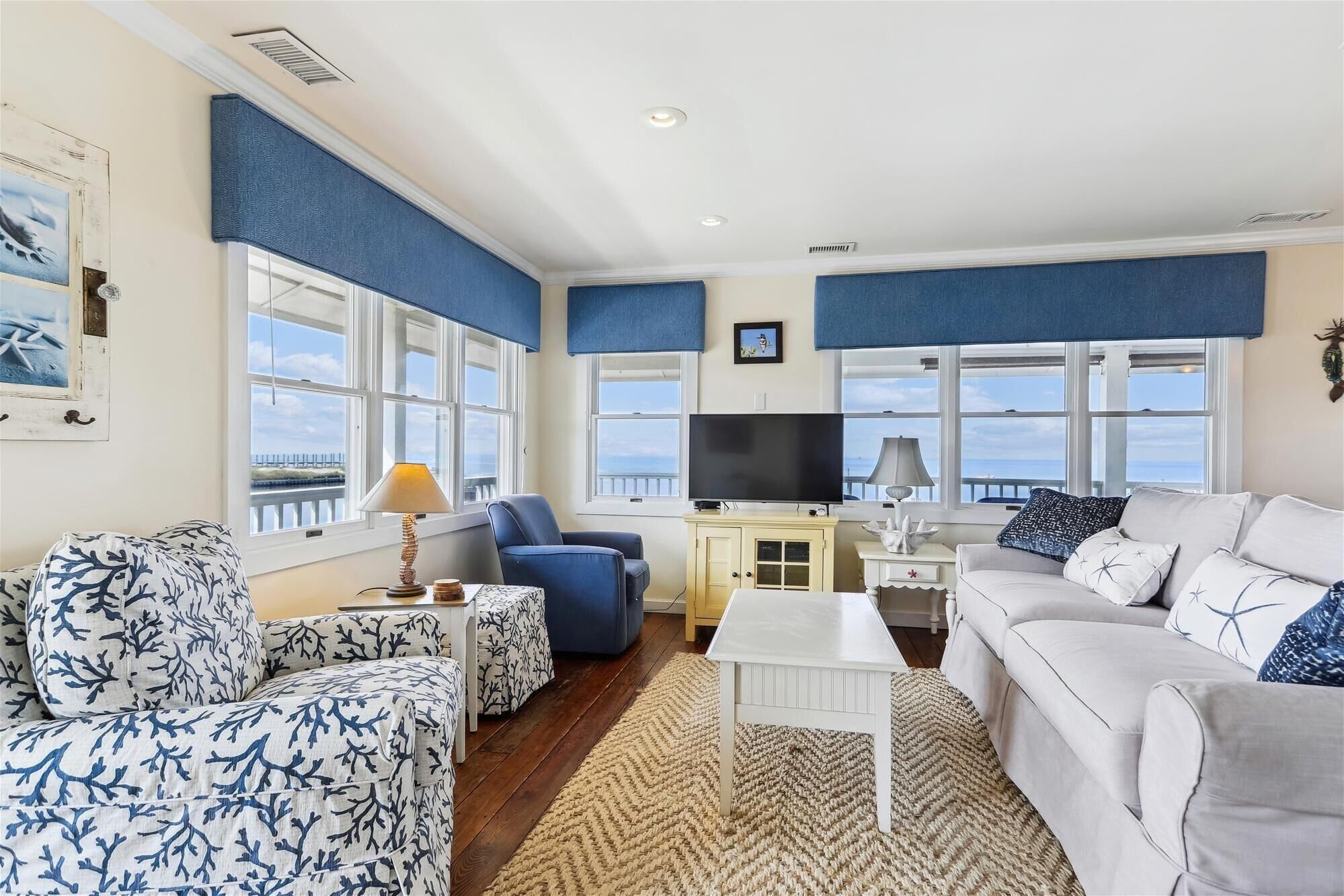 A living room with a view of the sea, featuring a sofa, chairs, a TV, and a small table.