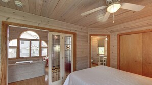 Dormitorio de cabaña de madera con una cama, un ventilador de techo y una ventana con vista a una casa.