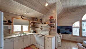 Cocina compacta con armarios de madera, estufa y microondas. Hay repisas sobre la encimera con vajilla y un reloj en la pared.