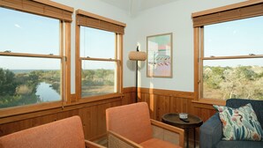 Ein Zimmer mit großen Fenstern, Holzwänden, einem gerahmten Kunstwerk und einem blauen Sofa mit einem gemusterten Kissen.