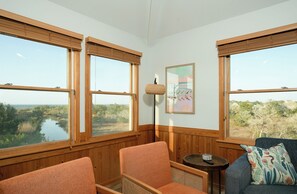 A room with large windows, wooden walls, a framed artwork, and a blue sofa with a patterned pillow.