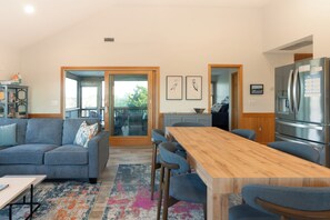 Cozinha moderna com mesa de jantar de madeira e banquetas, um sofá cinza, e vista para área externa através de uma porta de vidro.