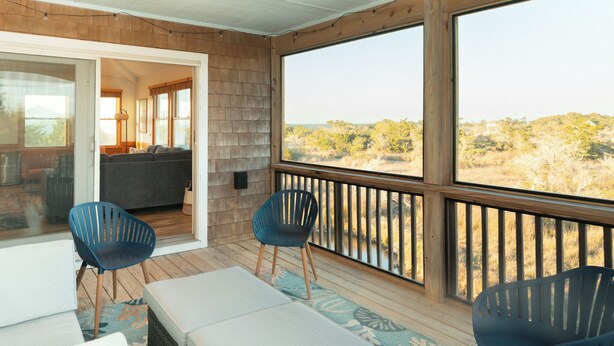 Ein überdachter Außenbereich mit Holzterrasse, zwei blauen Stühlen, einer Couch und Blick auf eine natürliche Landschaft.