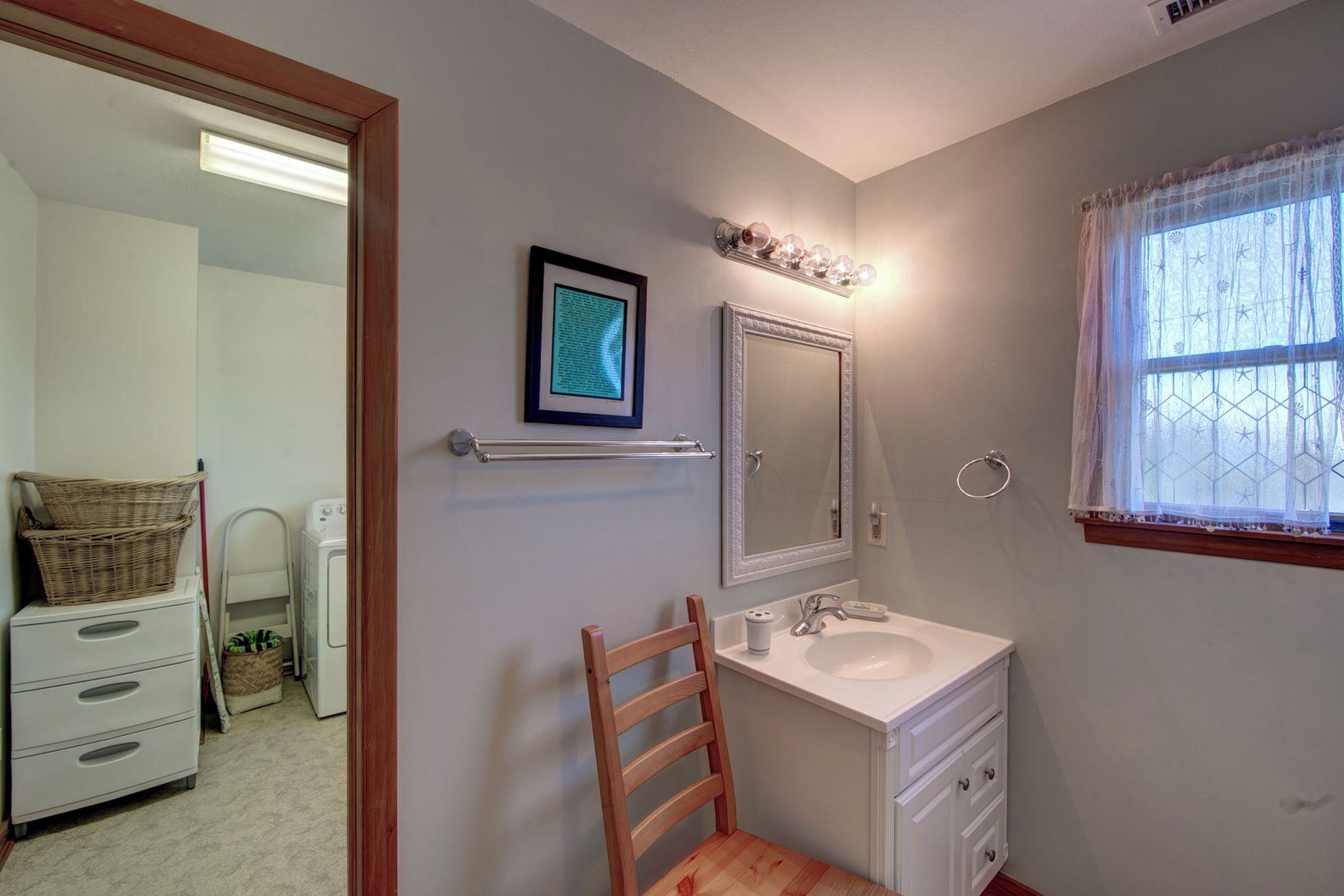 Une salle de bain avec un lavabo blanc, un miroir et un porte-serviettes. On y trouve une chaise en bois et un panier en osier posés sur une commode.
