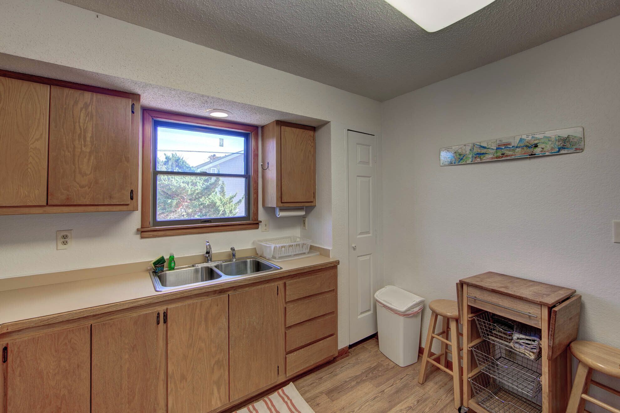 A kitchen with wooden cabinets, a double sink, a window, a trash bin, and a small stool.