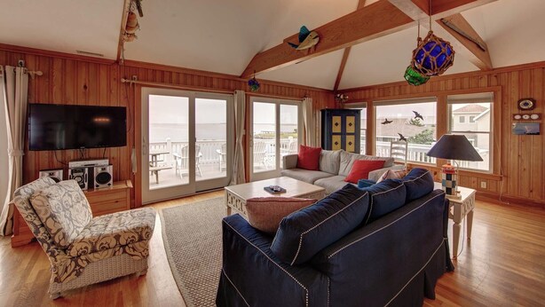 A spacious living room with a large TV, wooden furniture, and a balcony with outdoor seating. - Blue Oyster - Ocracoke Island (Ocracoke)