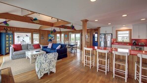 A spacious living area with a wooden floor, a blue sofa, a patterned ottoman, and a dining table with white chairs. - Blue Oyster - Ocracoke Island (Ocracoke)