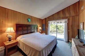 A bedroom with wooden paneling, a bed with a patterned bedspread, two bedside lamps, a small table with a lamp, a dresser, and a window with a view of greenery. - Shrimp and Grits - Ocracoke Island (Ocracoke)