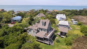 A large house with a prominent front porch and multiple levels, surrounded by lush greenery and a view of the ocean.
