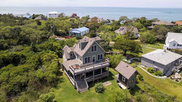 A large house with a deck and a smaller attached structure, surrounded by dense greenery and a view of the sea.