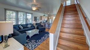 A spacious living room with a large sofa, a coffee table, and a blue rug. There is a wooden staircase leading to another level. - The Dory - Ocracoke Island (Ocracoke)