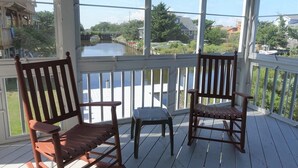 Una terraza de madera con dos mecedoras y una pequeña mesa, con vistas a un canal que tiene barcos y casas al fondo.