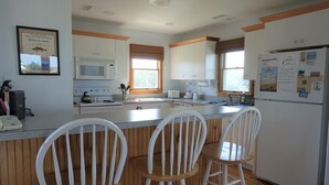 Una cocina con armarios blancos, un microondas, un refrigerador y una ventana. Hay tres taburetes blancos y una cafetera en la encimera.