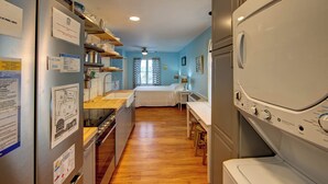 Une cuisine compacte équipée d’un four encastré, d’un évier et d’une cuisinière. On y trouve également un coin repas avec une table et des chaises. La pièce est dotée d’un plancher en bois et d’une fenêtre avec des rideaux.