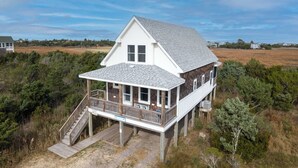 Une maison à deux étages dotée d’un porche couvert et d’une petite terrasse, entourée d’une végétation luxuriante et de vastes champs ouverts.