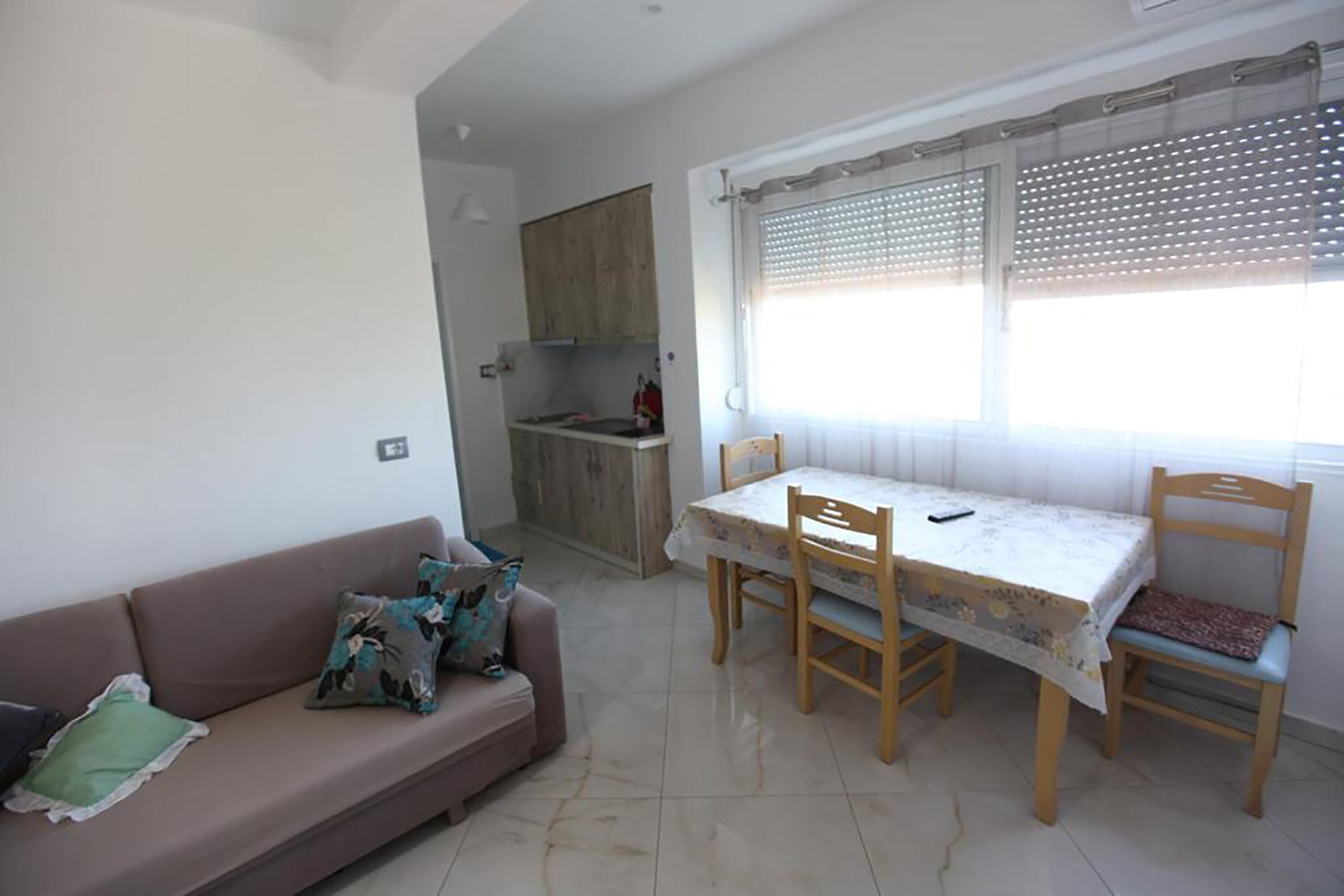 A living room with a sofa, a dining table with chairs, a kitchen area, and a window with blinds.