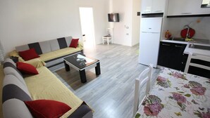 En moderne stue med sofa, sofabord og spisebord med stole. Der er et køkkenområde med hvide skabe og en sort bordplade.